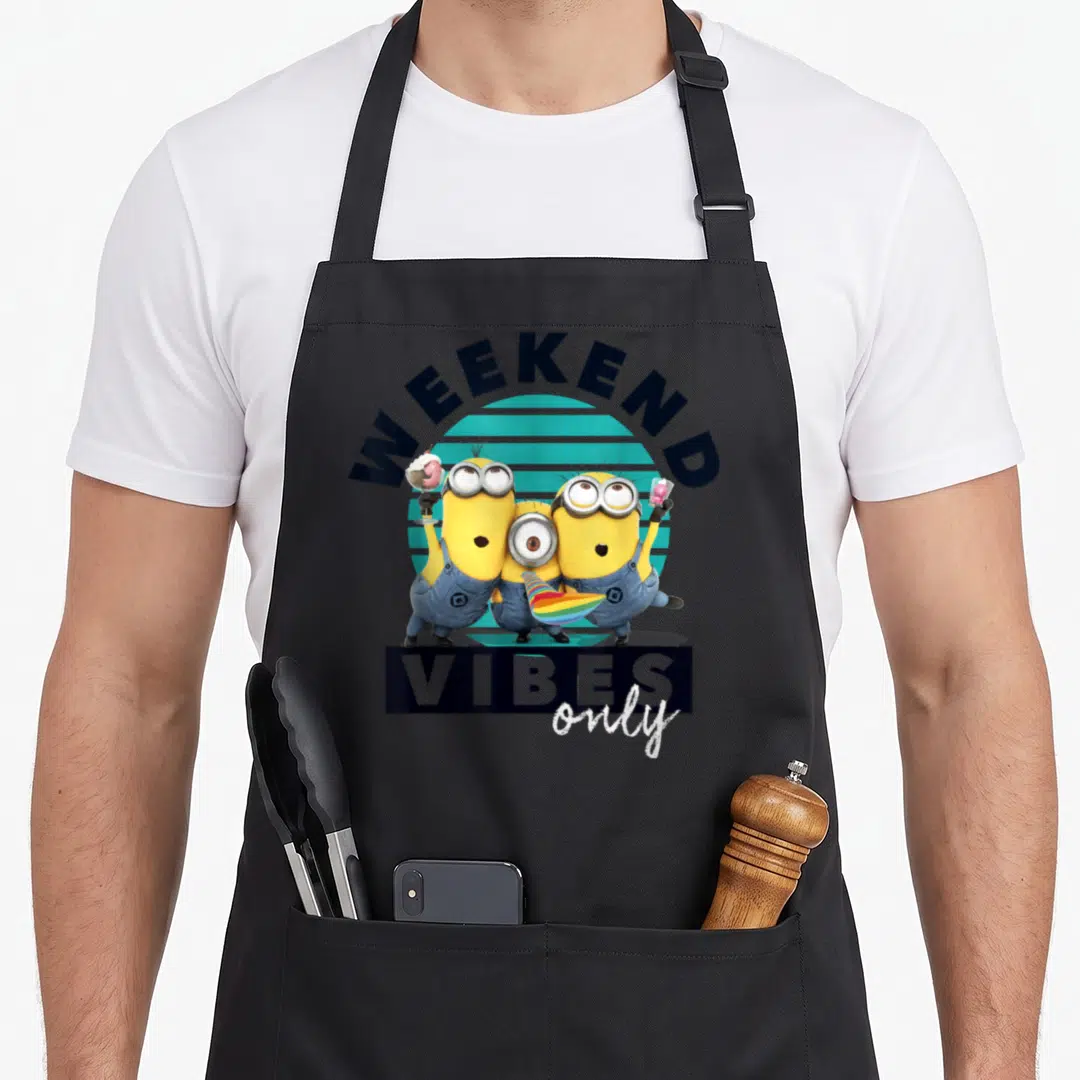 Despicable weekend vibes apron - Three yellow minions holding ice cream and a party horn under arched text over a teal — shown on a black cotton canvas apron against a white background
