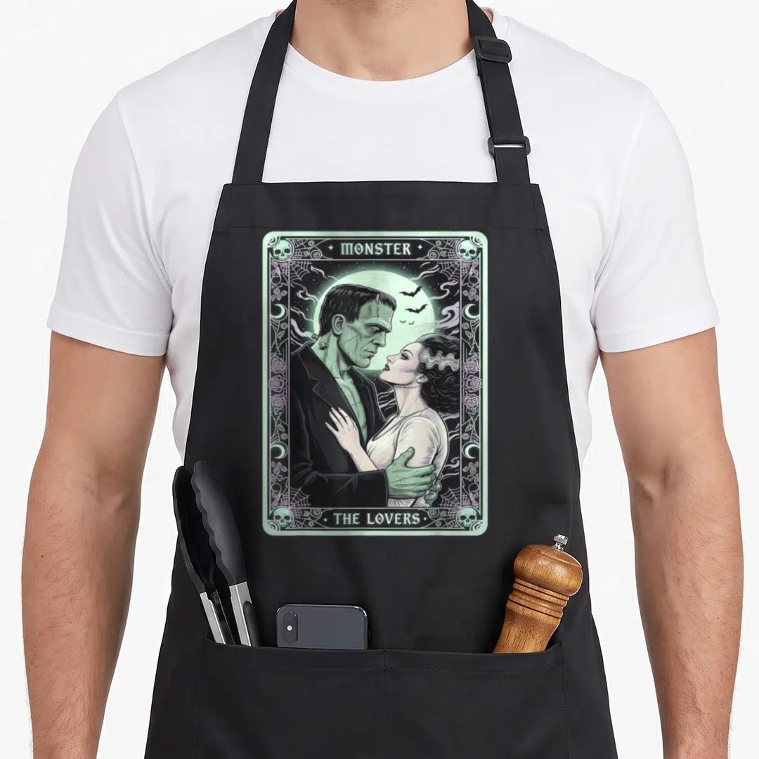 Card bride apron - Retro tarot card print of Frankenstein and bride embracing before a moon with skull border — shown on a black cotton canvas apron against a white background