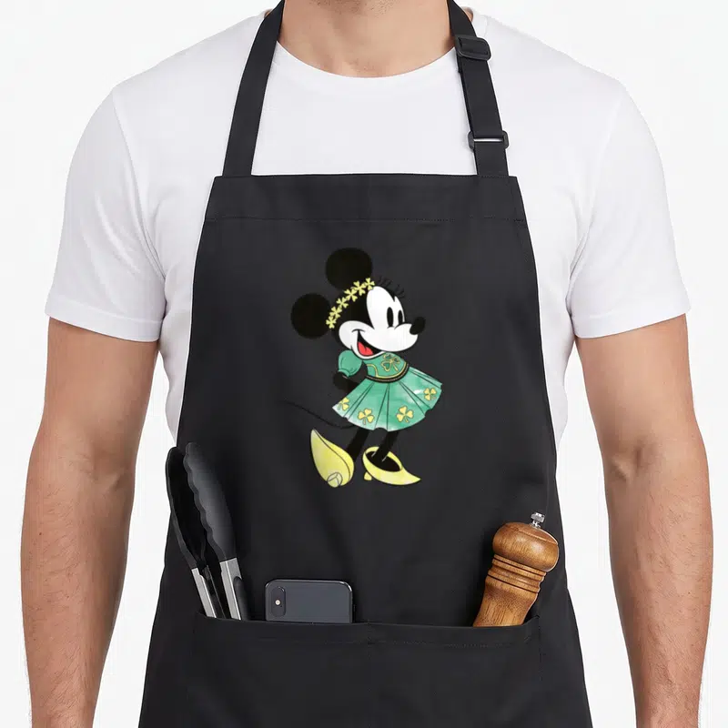 Irish st patrick day aprons - Cartoon Minnie Mouse in green dress with yellow shamrocks and flower headband — shown on a black cotton canvas apron against a white background