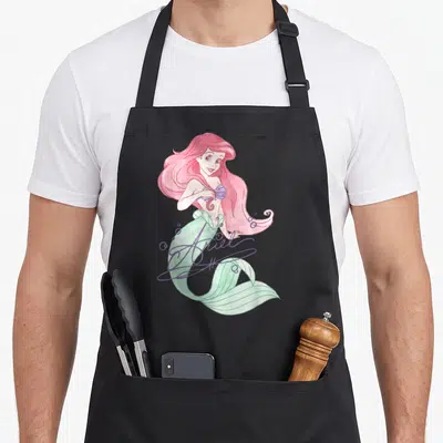 Signed mermaid apron - Watercolor Ariel mermaid with pink hair, green tail, and purple script signature — shown on a black cotton canvas apron against a white background