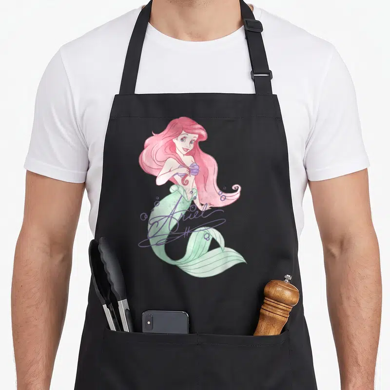 Signed mermaid apron - Watercolor Ariel mermaid with pink hair, green tail, and purple script signature — shown on a black cotton canvas apron against a white background