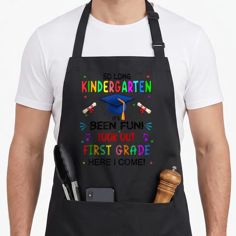 Class come and get it apron - Vertical rainbow text reading So Long Kindergarten Look Out First Grade with blue cap — shown on a black cotton canvas apron against a white background