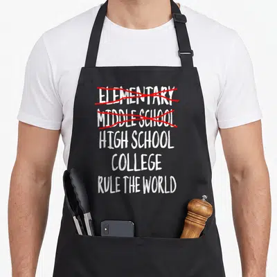 School junior apron - Stacked white text crossing out elementary and middle school with red scribbles above high school — shown on a black cotton canvas apron against a white background