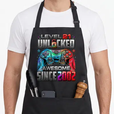 Unlocked 21st gaming aprons - Cartoon game controller with LEVEL 21 UNLOCKED and SINCE 2002 text in grey red blue — shown on a black cotton canvas apron against a white background