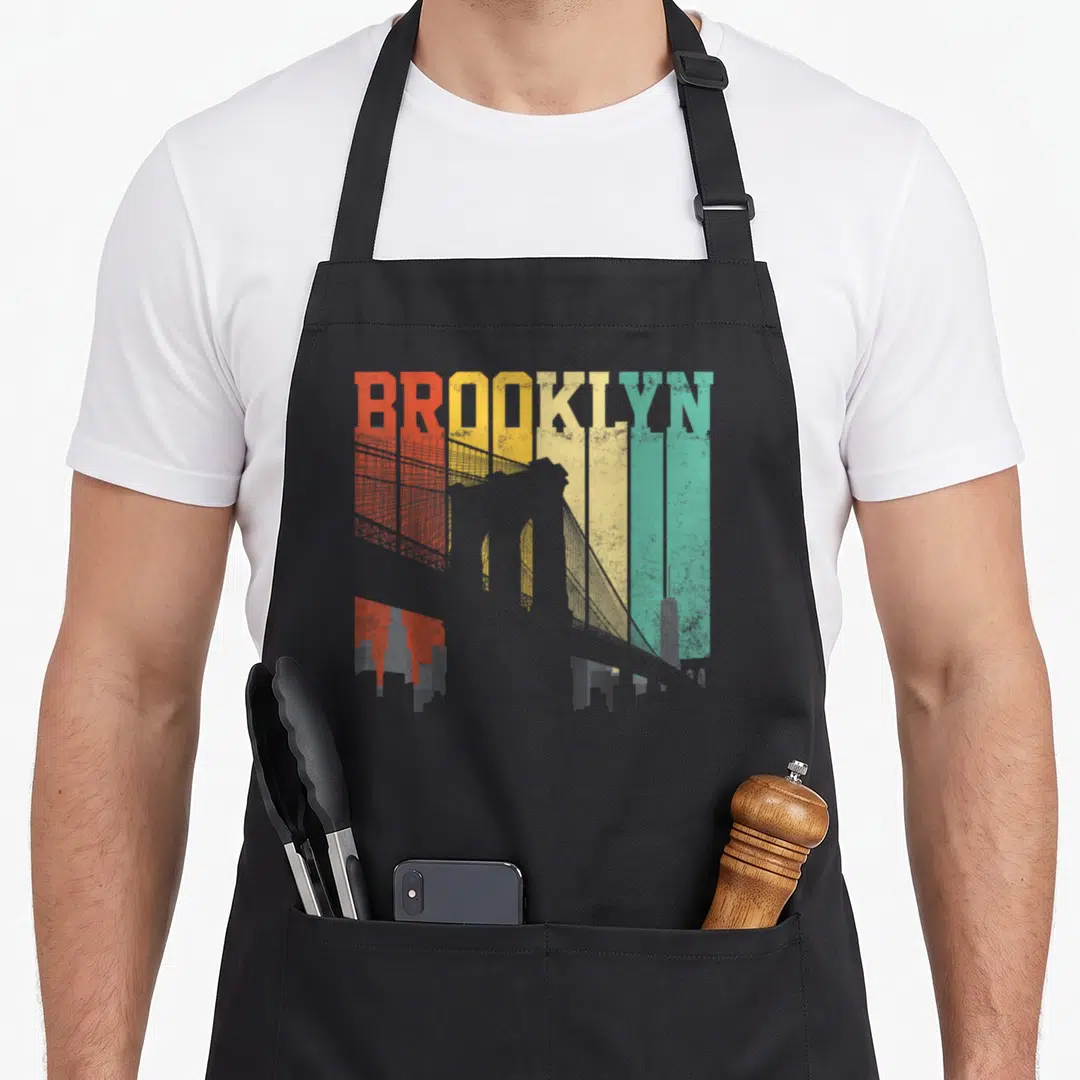 Brooklyn bridge new york apron - BROOKLYN text over grey bridge silhouette on red orange yellow and teal vertical — shown on a black cotton canvas apron against a white background