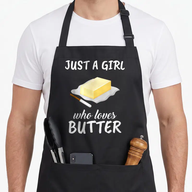 Clothe butter apron - Cartoon yellow butter block on foil with knife and white text Just A Girl Who Loves Butter — shown on a black cotton canvas apron against a white background