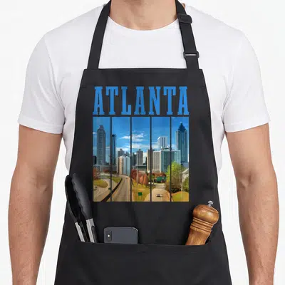 Atlanta pride apron - Blue ATLANTA text above five vertical panels of a grey and green city skyline in retro style — shown on a black cotton canvas apron against a white background