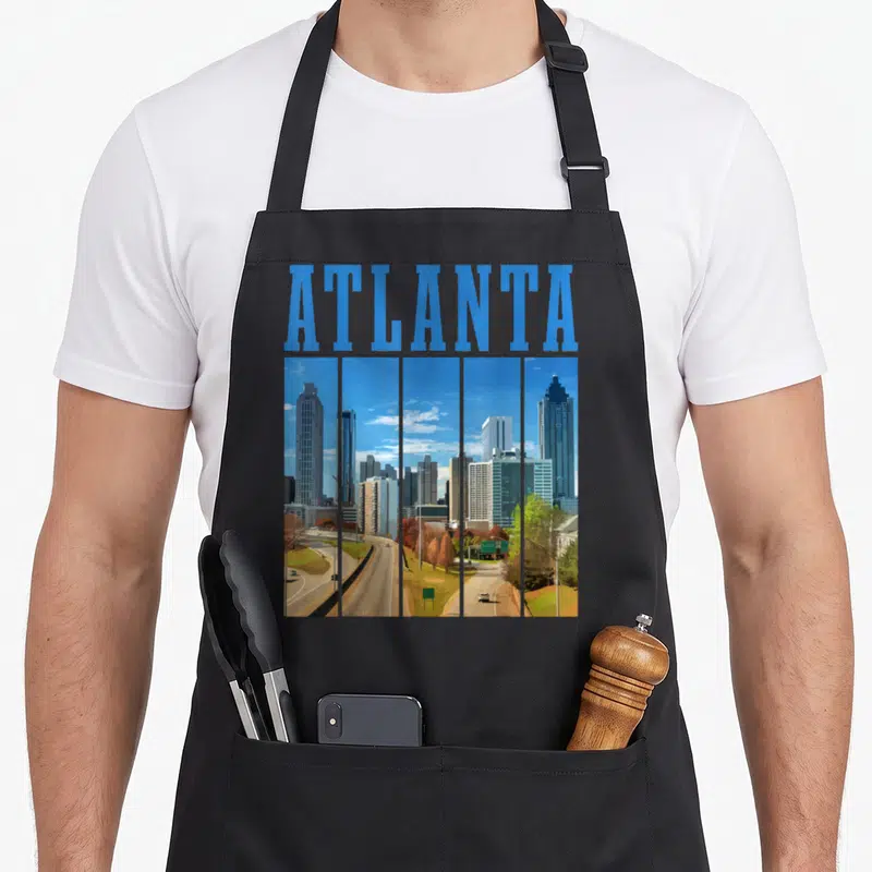 Atlanta pride apron - Blue ATLANTA text above five vertical panels of a grey and green city skyline in retro style — shown on a black cotton canvas apron against a white background
