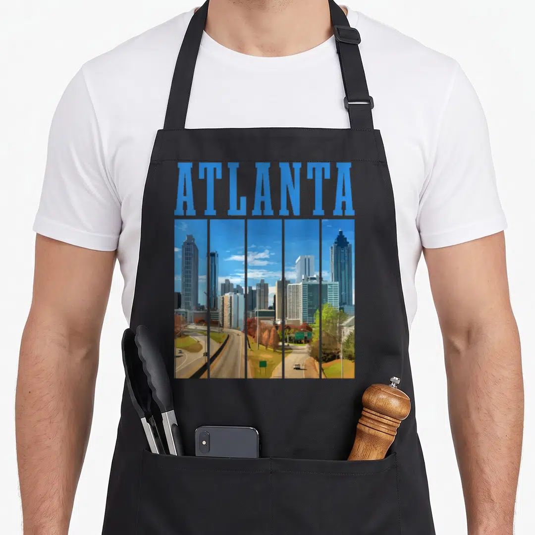 Atlanta pride apron - Blue ATLANTA text above five vertical panels of a grey and green city skyline in retro style — shown on a black cotton canvas apron against a white background