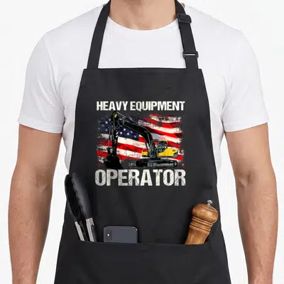 Heavy american flag apron - Yellow excavator on distressed American flag with HEAVY EQUIPMENT OPERATOR text — shown on a black cotton canvas apron against a white background
