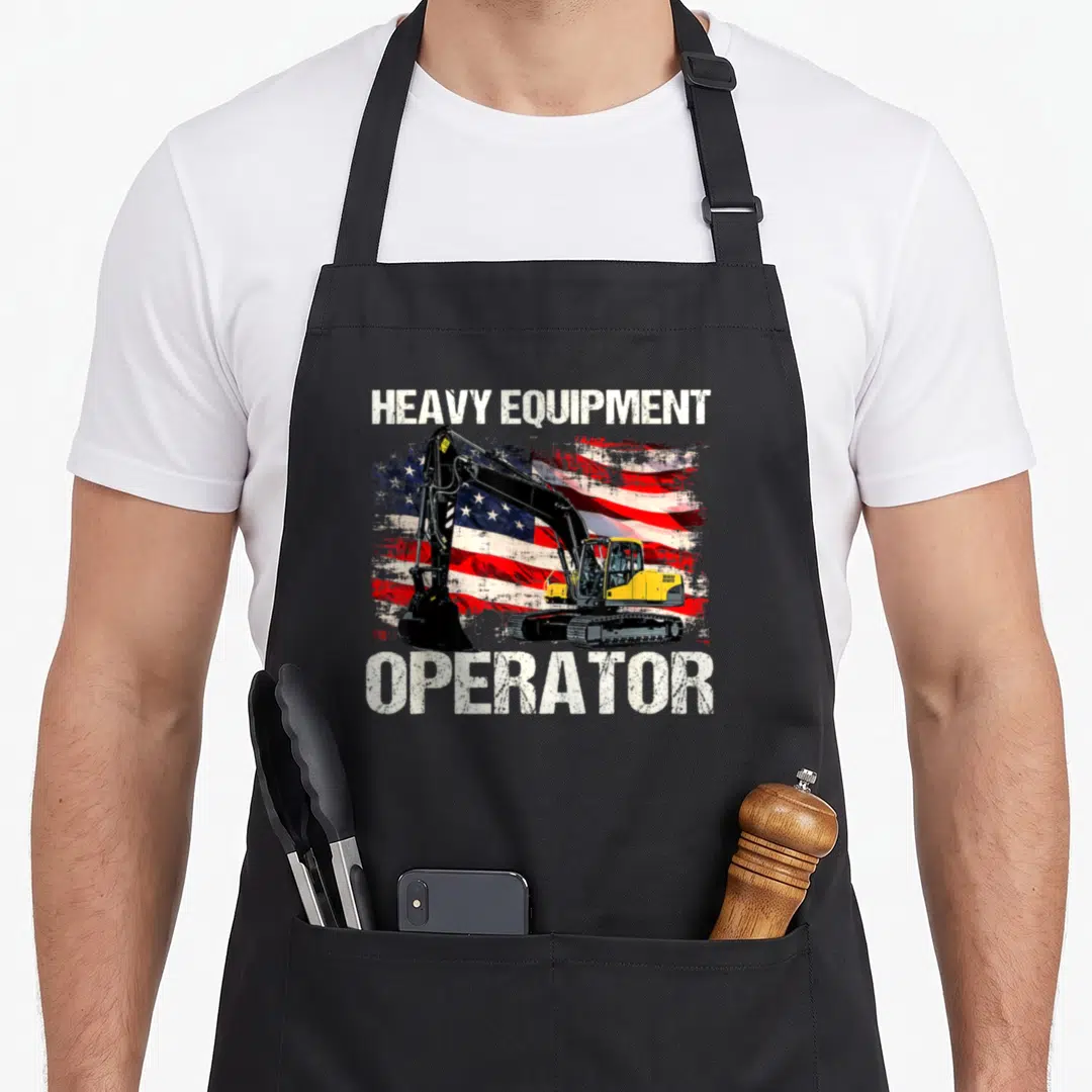 Heavy american flag apron - Yellow excavator on distressed American flag with HEAVY EQUIPMENT OPERATOR text — shown on a black cotton canvas apron against a white background