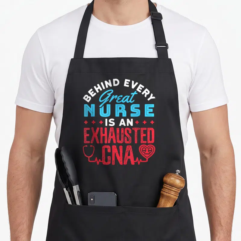 Exhausted nursing aprons - Stacked text reading Behind Every Great Nurse Is An Exhausted CNA with red stethoscope — shown on a black cotton canvas apron against a white background