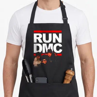 Run outline of an apron - Stacked RUN DMC text in white and red with three portraits overlapping the bottom bar — shown on a black cotton canvas apron against a white background