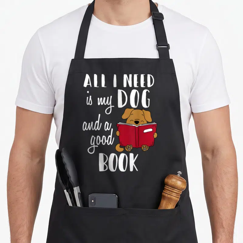 Funny book dog apron - Cartoon brown dog holding open red book surrounded by white text — shown on a black cotton canvas apron against a white background