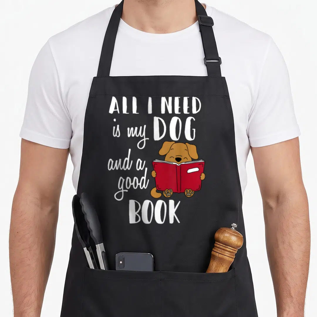 Funny book dog apron - Cartoon brown dog holding open red book surrounded by white text — shown on a black cotton canvas apron against a white background