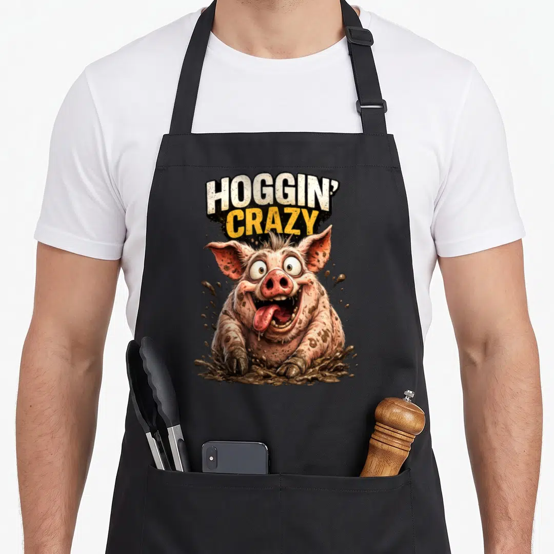 Hoggin crazy pig lady apron - Cartoon pig sitting in brown mud splash below stacked HOGGIN' CRAZY text in white — shown on a black cotton canvas apron against a white background