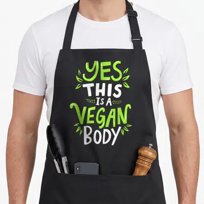 Natural vegan apron - Stacked yes this is a vegan body text with green leaves flanking lines — shown on a black cotton canvas apron against a white background