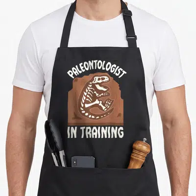 Paleontologist training apron - White dinosaur skeleton in brown rock with arched text — shown on a black cotton canvas apron against a white background