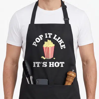 Pop popcorn apron - Minimalist striped popcorn bucket with POP IT LIKE IT'S HOT text in white red and yellow — shown on a black cotton canvas apron against a white background