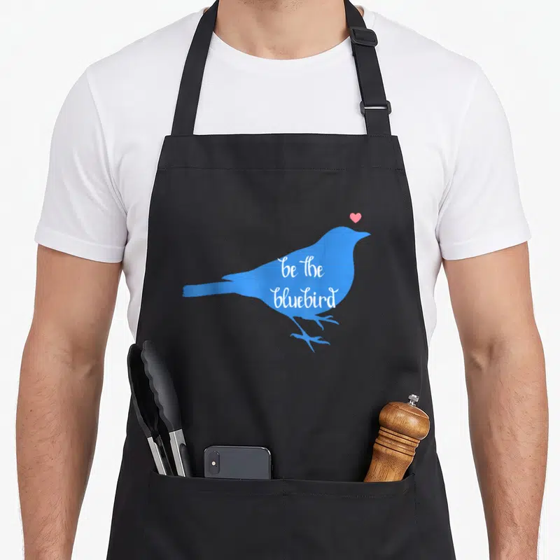 Happiness bluebird aprons - Blue bird silhouette with pink heart and white be the bluebird text — shown on a black cotton canvas apron against a white background