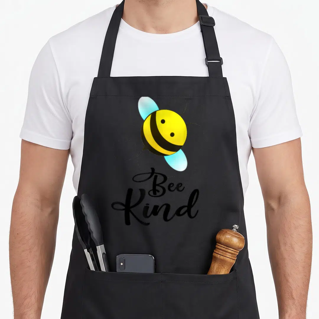 Funny kind bee apron - Cartoon bee with blue wings and dotted trail above Bee Kind text in black script — shown on a black cotton canvas apron against a white background