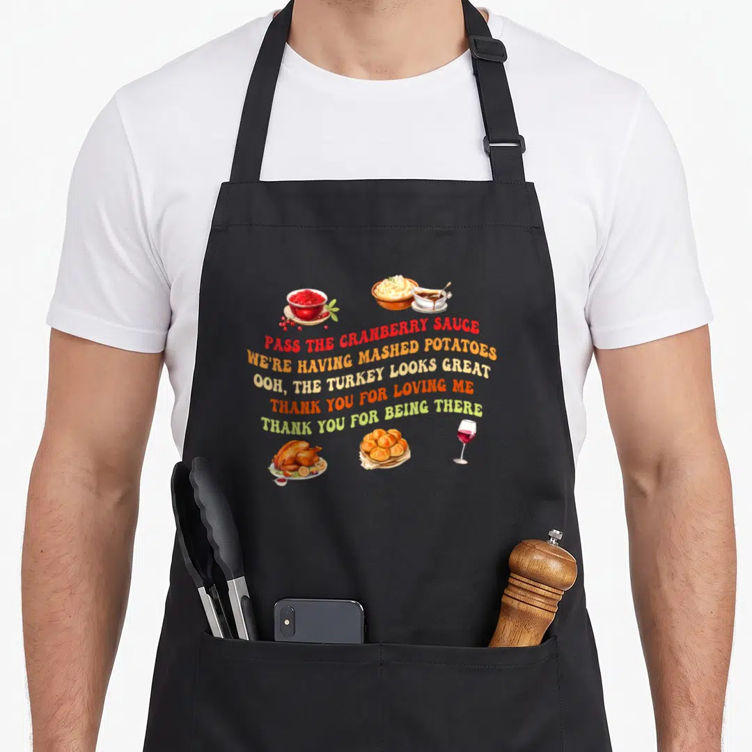 Sauce mashed apron - Arched text reading Pass The Cranberry Sauce above cartoon turkey, rolls, and wine in red orange — shown on a black cotton canvas apron against a white background
