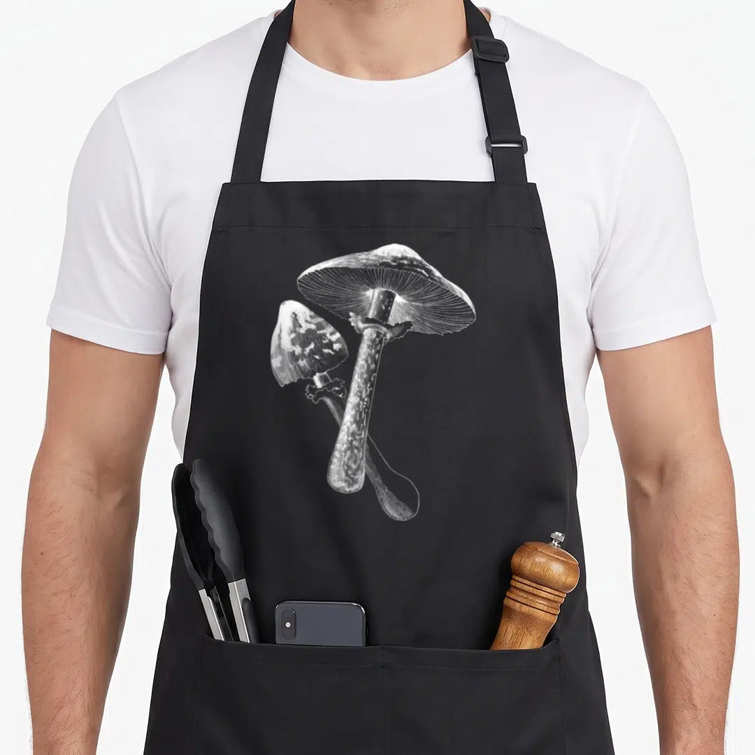 Whisperer foraging apron - Two white mushrooms in line art, one large overlapping a smaller one — shown on a black cotton canvas apron against a white background