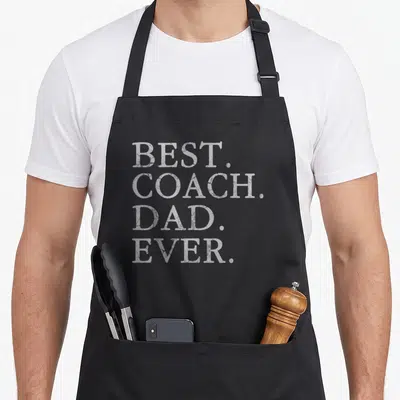 Hockey football apron - White distressed text reading BEST. COACH. DAD. EVER. stacked on four lines — shown on a black cotton canvas apron against a white background