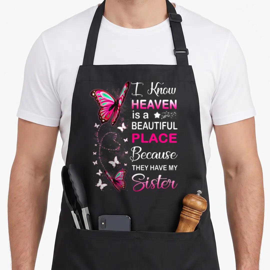 Sister angels and aprons - Pink and teal butterflies with text about a sister in heaven on a black background — shown on a black cotton canvas apron against a white background