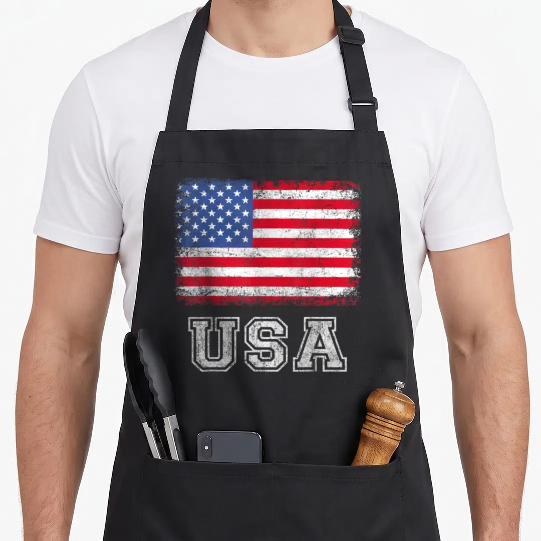 Funny patriotic american flag apron - Distressed red white blue flag above white USA block letters — shown on a black cotton canvas apron against a white background