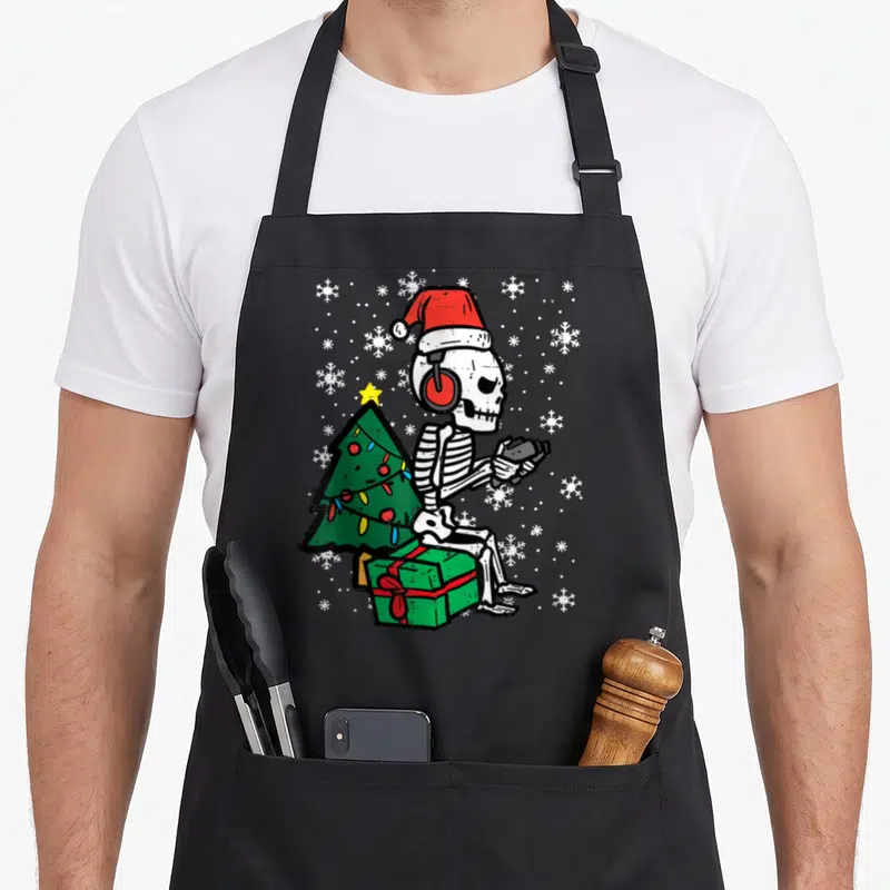 Gamer christmas santa apron - Cartoon skeleton with red headphones sitting on green gift near Christmas tree — shown on a black cotton canvas apron against a white background