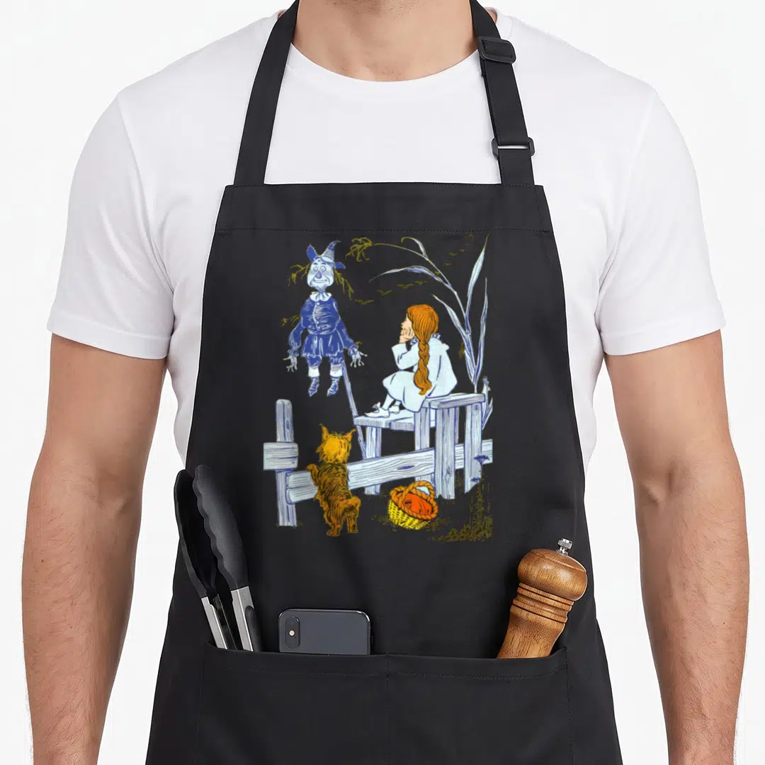 Toto dorothy apron - Line art of scarecrow, girl on fence, dog, and basket in blue orange white yellow — shown on a black cotton canvas apron against a white background