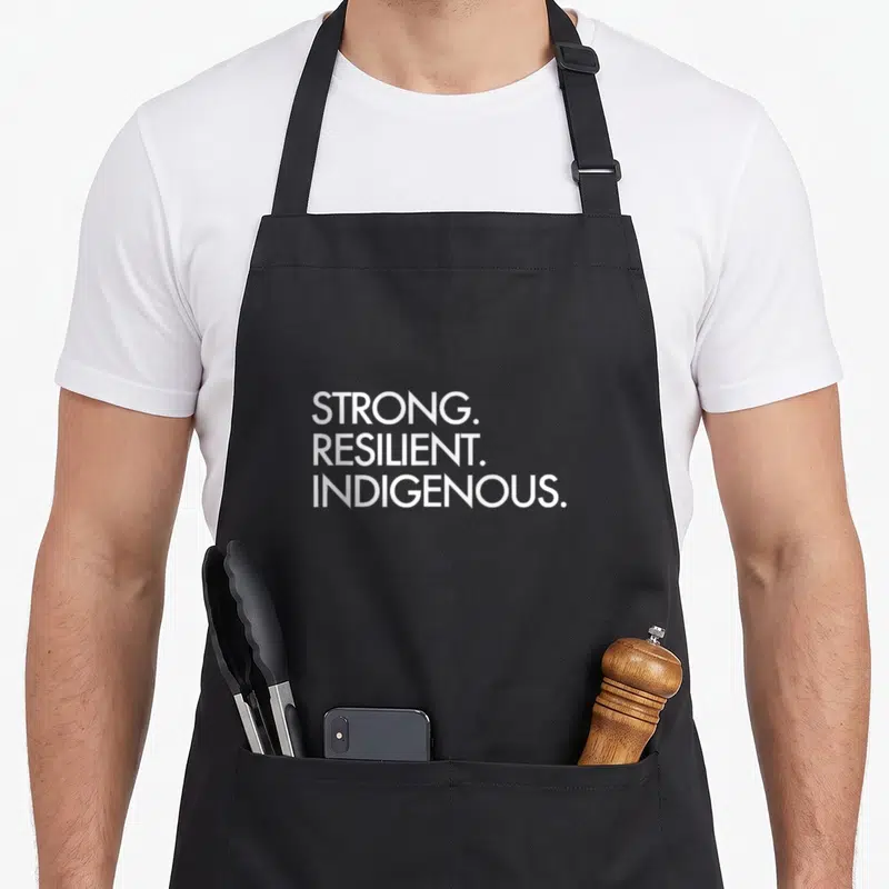 Duty heavy apron - White stacked text STRONG RESILIENT INDIGENOUS on black fabric — shown on a black cotton canvas apron against a white background