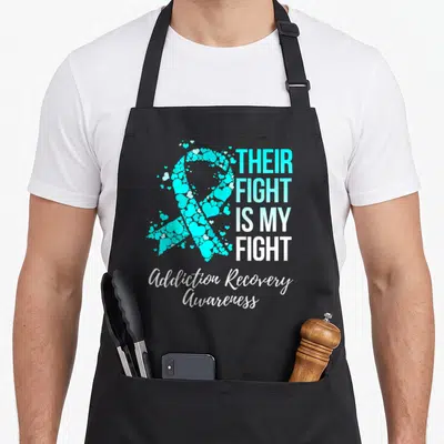 Their addiction recovery apron - Cyan and white heart ribbon formed by small hearts with stacked text and script footer — shown on a black cotton canvas apron against a white background