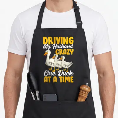 Husband duck apron - Three cartoon ducks with yellow beaks and text Driving My Husband Crazy One Duck At A Time — shown on a black cotton canvas apron against a white background