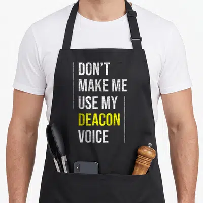 Use catholic apron - Stacked white and yellow text reading DON'T MAKE ME USE MY DEACON VOICE framed by vertical lines — shown on a black cotton canvas apron against a white background