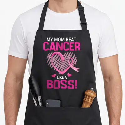 Beat boss apron - Pink and white text My Mom Beat Cancer Like A Boss surrounding a heart-shaped awareness ribbon — shown on a black cotton canvas apron against a white background
