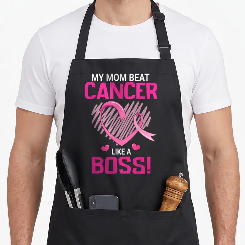Beat boss apron - Pink and white text My Mom Beat Cancer Like A Boss surrounding a heart-shaped awareness ribbon — shown on a black cotton canvas apron against a white background