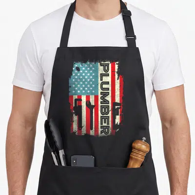 Plumber 4th of july apron - Vertical PLUMBER text over distressed USA flag with pipe wrench, wrench, and plunger — shown on a black cotton canvas apron against a white background