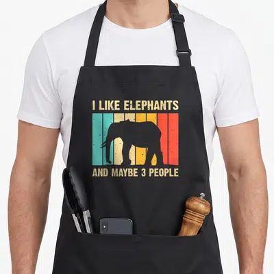 Elephant print apron for dad - Grey elephant silhouette on multicolored stripes with beige text reading I like — shown on a black cotton canvas apron against a white background
