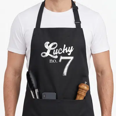 Lucky distressed apron - White Lucky no. 7 typography with distressed texture on dark background — shown on a black cotton canvas apron against a white background