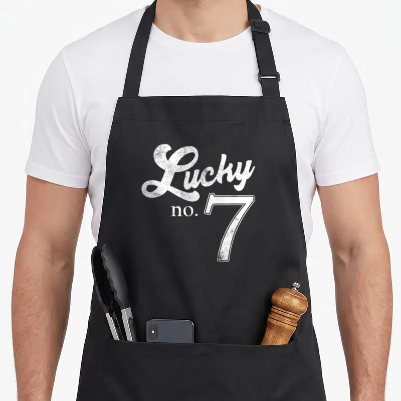 Lucky distressed apron - White Lucky no. 7 typography with distressed texture on dark background — shown on a black cotton canvas apron against a white background