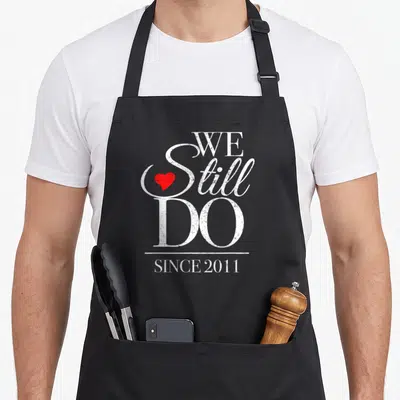 15th distillery apron - Stacked white text We Still Do Since 2011 with small red heart inside S and horizontal line — shown on a black cotton canvas apron against a white background