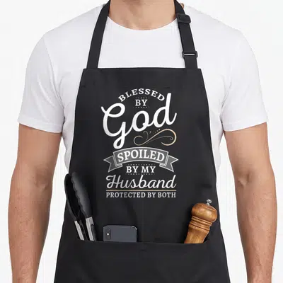 Spoiled husband apron - White and tan stacked text reading Blessed by God Spoiled by My Husband Protected by Both — shown on a black cotton canvas apron against a white background