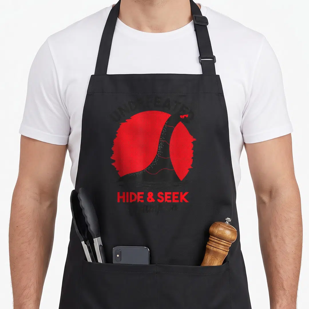 Loch undefeated apron - Nessie monster head in red halftone circle with UNDEFEATED HIDE & SEEK Champion text — shown on a black cotton canvas apron against a white background