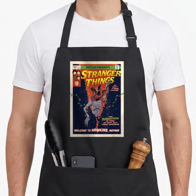 Welcome cover apron - Retro comic cover with Demogorgon upside down and yellow Stranger Things title text — shown on a black cotton canvas apron against a white background