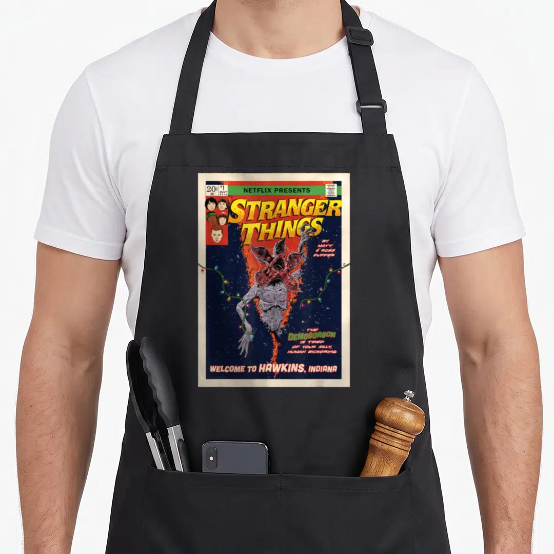 Welcome cover apron - Retro comic cover with Demogorgon upside down and yellow Stranger Things title text — shown on a black cotton canvas apron against a white background