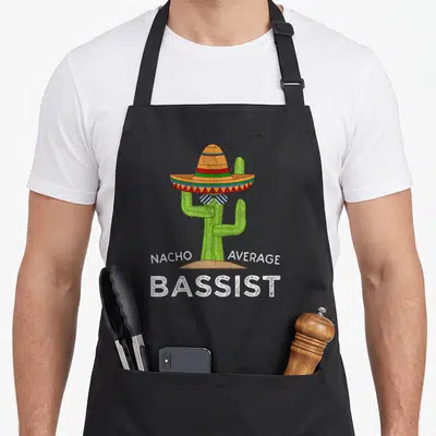 Guitar player bassist apron - Cartoon green cactus in orange sombrero with white text reading NACHO AVERAGE BASSIST — shown on a black cotton canvas apron against a white background