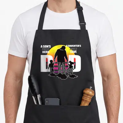 Hero father day apron - DAD text with father and children silhouettes against yellow sun and A Son's First Hero — shown on a black cotton canvas apron against a white background