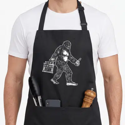 Bigfoot craft apron - White line-art bigfoot holding beer carrier and bottle — shown on a black cotton canvas apron against a white background